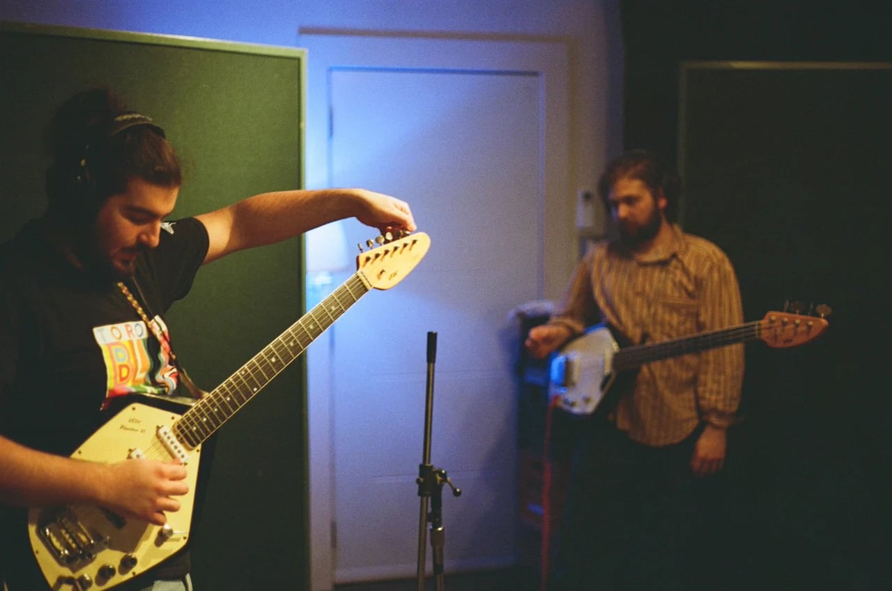 Two musicians playing bass and guitar during a recording session under green and blue studio lighting.