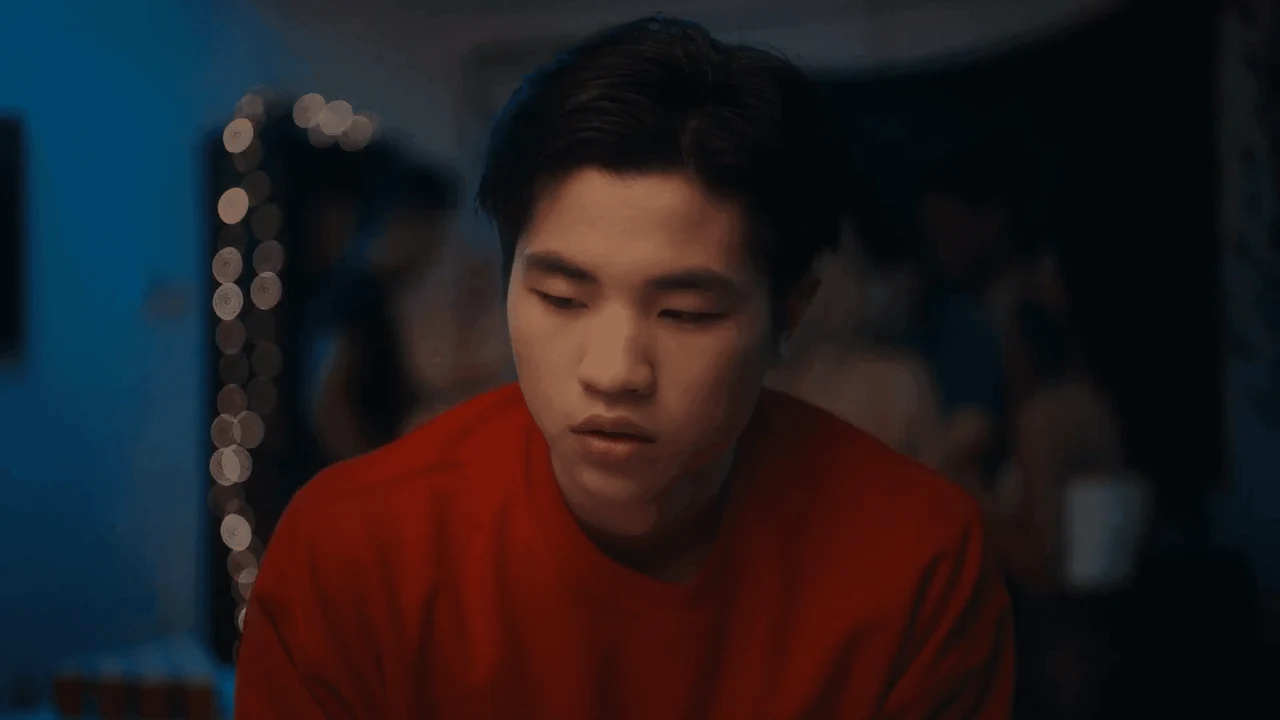 A young man in a red shirt in a dimly lit scene from Tiger Mom.